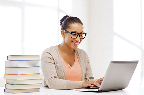A young woman sitting a desk with laptop open and looking at screen and smiling. There is a stack of textbooks next to her.